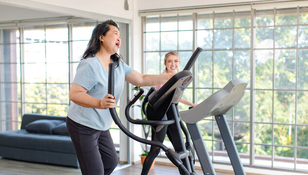 Asian happy healthy old chubby retirement pensioner mother smiling using elliptical machine exercise working out with young beautiful daughter runner walking running on adjustable pad in living room