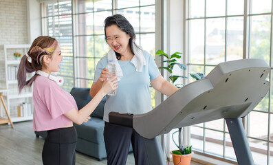 Asian old chubby retirement pensioner happy healthy female sport fitness woman in casual sportswear walking running on machine treadmill while young trainer bringing giving water bottle for resting