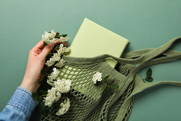 Book in string bag, flowers and female hand on green background, top view