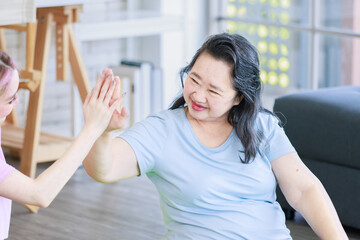 Fototapeta premium Asian old senior pensioner retirement cheerful happy healthy fit mother in casual sportswear and young beautiful daughter holding raising hand high five watching doing yoga stretching in living room.