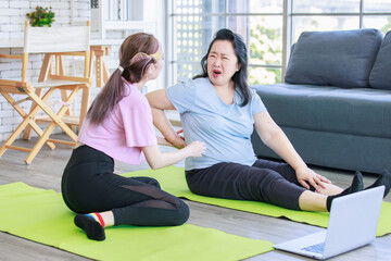 Asian old senior chubby pensioner retirement mother sitting on yoga mat having backache from stretching via online lesson when young daughter helping supporting checking massaging back in living room