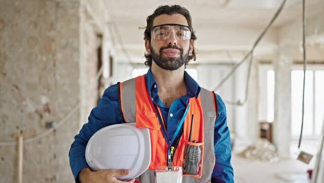 Young hispanic man architect standing with serious face holding hardhat at construction site