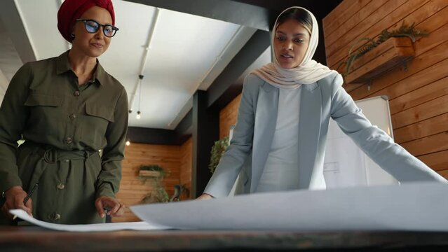Female designers in headscarves discussing blueprint drawings as they take on an architectural project. Two architects contributing their expertise to the designing of a floor plan in an office.