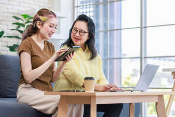 Asian young pretty daughter and old senior pensioner retirement cheerful happy mother sitting on sofa couch in living room at home together surfing browsing internet with tablet and laptop computer