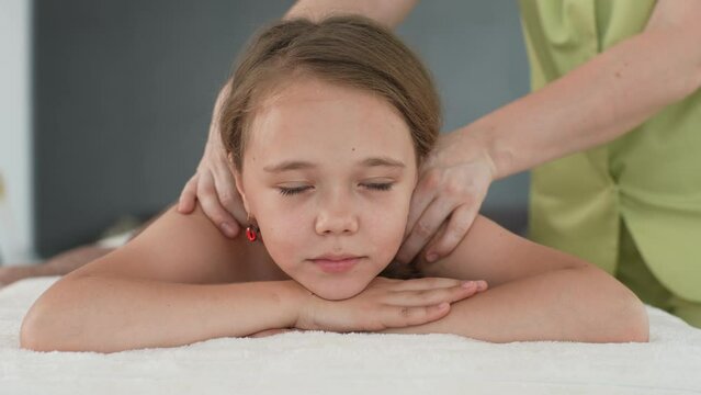 Portrait of cheerful little girl having neck, shoulder and back massage by unrecognizable female pediatric masseuse at medical clinic. Adorable preteen kid feeling happy and relax during back massage.