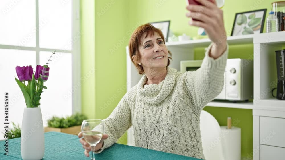 Wall mural Mature hispanic woman doing a video call sitting on table drinking white wine at dinning room