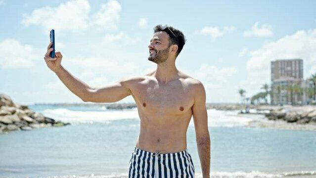 Young hispanic man tourist wearing swimsuit make selfie by smartphone at the beach