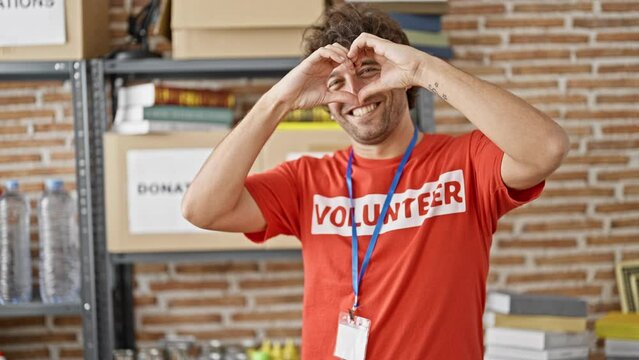 Young hispanic man activist wearing volunteer uniform doing heart gesture at charity center