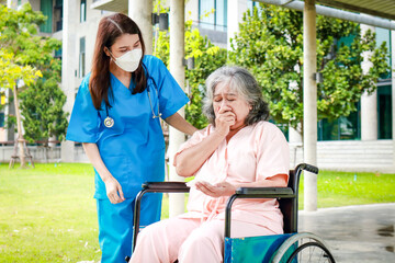 Asian female doctor in surgical gown caring for an elderly female patient sitting in a wheelchair outside a building. medical services in hospitals