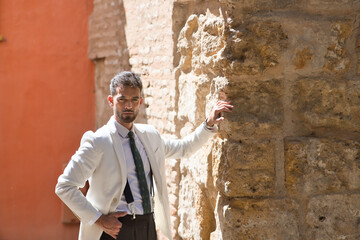 Fototapeta na wymiar Attractive young businessman with beard, suit and tie, posing with hand in pocket leaning against a stone wall looking at camera. Concept beauty, fashion, success, achiever, trend.