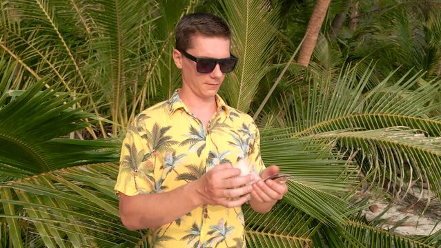 A young man in black sunglasses and a yellow tropical shirt counting Thai Baht with palm fronds in the background, on a sunny day in Thailand.