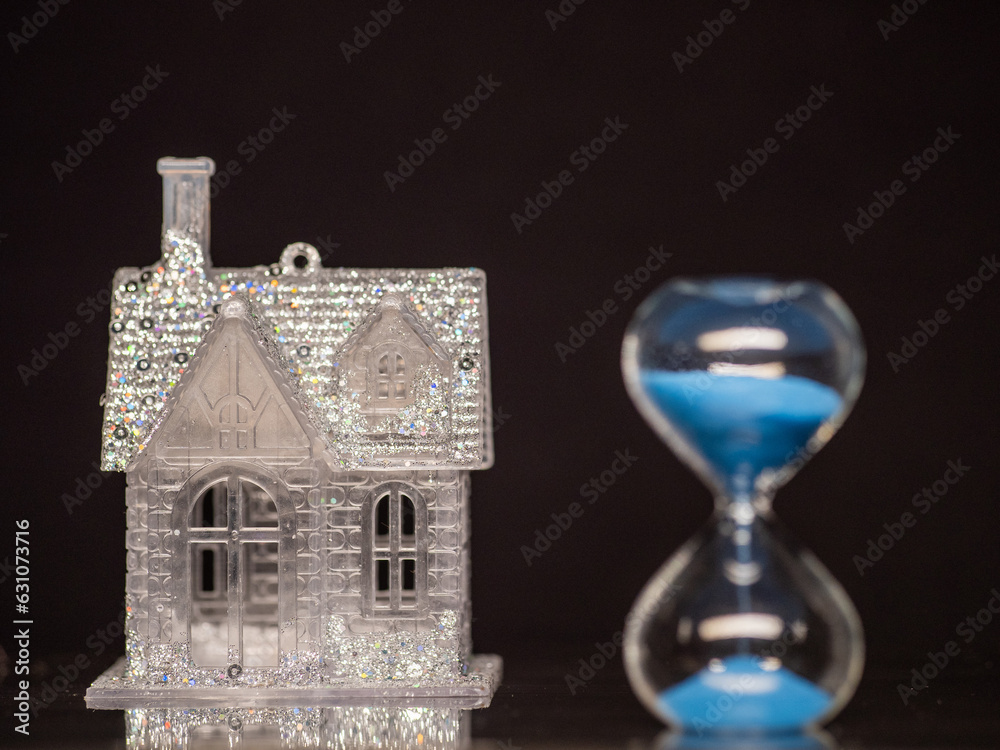 Wall mural Small glamourous house with glitter in focus, sand clock with blue sand out of focus. Dark background. Time to buy house concept. Invest in real estate theme. Banking and finance industry background