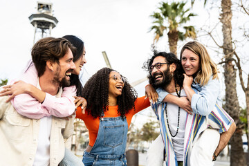 Multiracial group of friends giving piggyback ride and looking each other. Diverse young women and men having fun laughing and carefree staring at each other outside.
