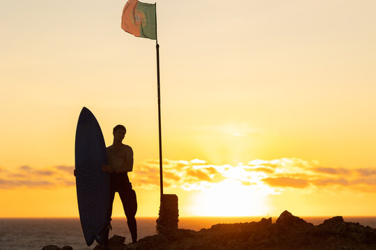 Silhouette of a man standing by the ocean at sunset holding a surfboard - the flag of Portugal on the flagpole - Powered by Adobe