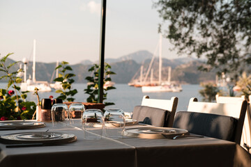 Fototapeta na wymiar served table in coast seaside restaurant, white and blue colors, sunset time, dinner at seaview with sailing boats at anchor