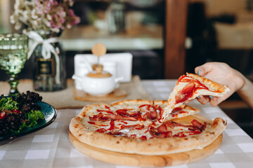 A slice of pizza with prosciutto, tomatoes, mozzarella and capers in hand over a wooden board on a...