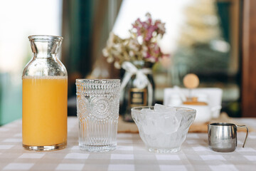 Decanter with orange juice next to decorative glass, glass plate with ice and metal mug on background of beige checkered tablecloth