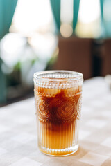 A mix of orange juice, coffee and ice in a decorative glass standing on a beige checkered tablecloth on a table on a blurred background