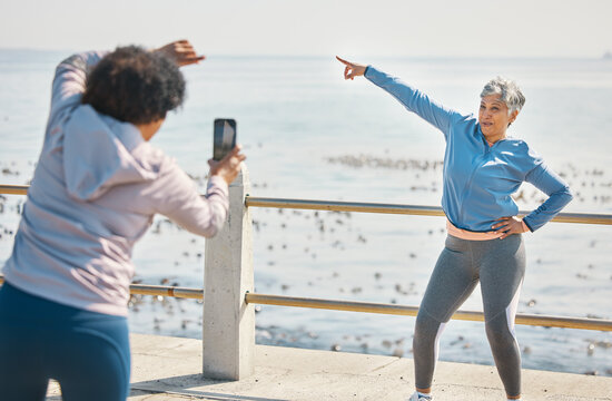 Phone, friends and photo of senior woman pointing at beach with silly pose at sea for fitness. Exercise, mobile and picture for social media post on a ocean promenade walk for workout and friendship