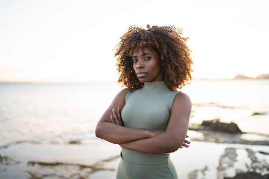 Serious African american young woman in dress looking at camera while standing against blurred beach at sunset