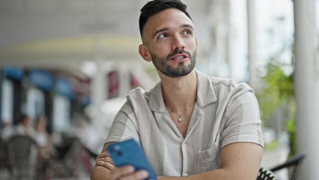 Young hispanic man using smartphone sitting on table speaking at coffee shop terrace