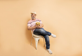 Retired senior indian man casual cloths and hat drinking coconut water while sitting on chair relaxing isolated on beige studio background. Hot summer season, Retirement life, Copy space. Full length