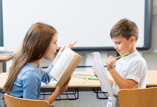Schoolchildren, a girl and a boy sitting at their desks in class and having a lively conversation. The schoolgirl explains the book to the boy, reads and tells. Schoolboy writing with interest and joy