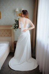 Female in wedding dress with strings at rear stand in room with flowers in hand. Bride hold arm on back and look aside.