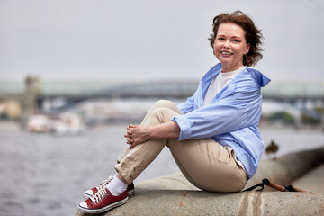 Fototapeta na wymiar Attractive smiling mature woman in casual clothes sits on the parapet of the river embankment. Senior lady relaxing outside. Middle aged brunette female smiles happily. Active lifestyle for elderly.