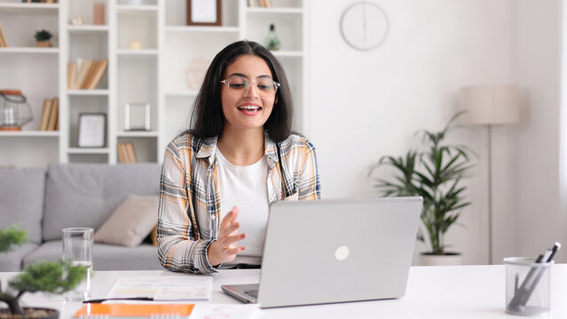 A young Indian girl works at a laptop and looks at the monitor. Beautiful woman talking on video call smiling. Studying in quarantine, listening to a podcast.