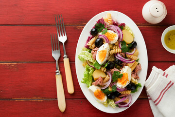 Tuna salad with pasta, eggs, potatoes, olives, red onions and sauce in white plate on old red rustic table background. Nicoise salad. French cuisine. Top view. Flat lay.
