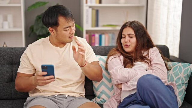 Man and woman couple using smartphone sitting on sofa arguing at home