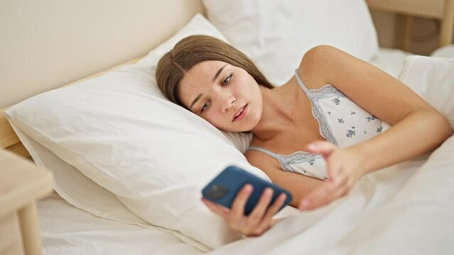 Young beautiful girl using smartphone lying on bed looking upset at bedroom