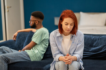 Unhappy young interracial couple having family problems, sitting on sofa at home, avoiding...