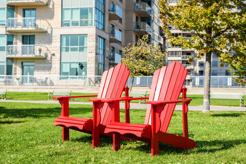 Red Adirondack chairs on a grass in a park o na sunny autumn day. Apartment buildings are visible...