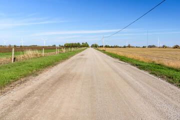 Straight gravel country road through farmland dotted with wind turbines on a clear autumn day