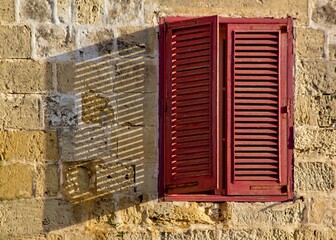 Red wooden louvered window with a shadow cast across the frame from the sunlight outside.