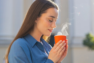 Young woman enjoying morning aroma coffee steam or tea hot drink outdoors. Relaxing, taking a break. Lovely girl walking in urban city sunny street sun rays, drinking coffee to go. Town lifestyles