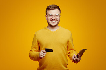 Young bearded man in eyeglasses wears yellow sweater, holds credit card and smartphone isolated on orange background. Online shopping. Online payment.