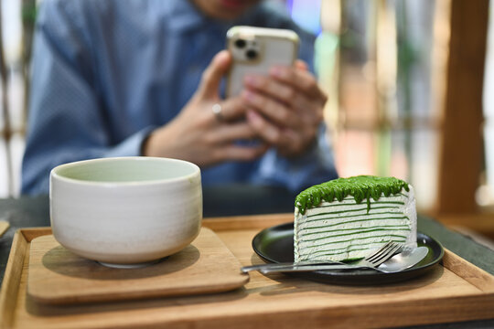 Close up and cropped image of Blogger using smartphone snap a picture of slice of Matcha crepe cake with Genmaicha Uji latte in the Japanese tradition ceramic bowl.
