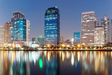 Night skyline of modern skyscrapers by lakeside, with glass curtain walls and dazzling city lights reflected in the smooth lake water in beautiful Benjakiti Park at blue dusk, in Bangkok, Thailand