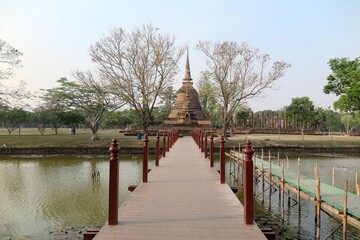 Fototapeta premium Scenery of Wat Sa Si, an ancient Buddhist Temple in Sukhothai Historical Park, with the majestic Pagoda (Stupa) in background & a wooden bridge across the canal ~ A beautiful heritage site in Thailand
