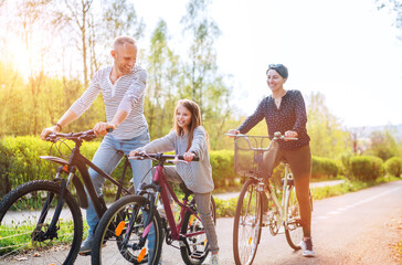Smiling father and mother with daughter during summer outdoor bicycle riding. They enjoy togetherness in the summer city park. Happy parenthood and childhood or active sport life concept image.