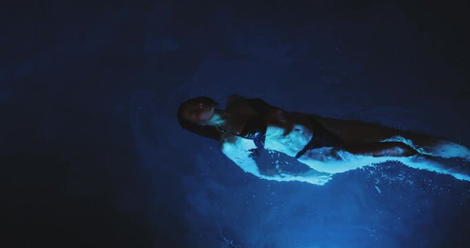 Top view og beautiful young adult sexy woman in a black bikini relaxing in the spa swimming pool. The fashionable model with perfect body swimming at night. Cinematic creative slow-motion 4k footage.