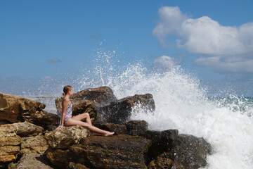a teenage girl in a bathing suit sits on a stone on the shore under sea spray