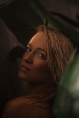 Attractive blonde woman portrayed in a close-up portrait, posing amidst palm leaves in a tropical shower setting