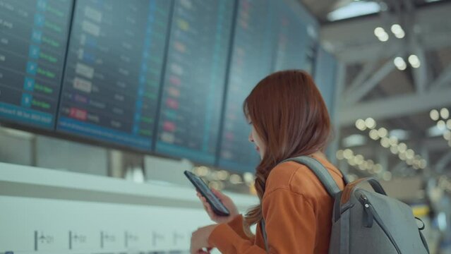 Happy asian woman traveller checking flight schedule departures board in airport terminal hall in front of check in counters. Tourist journey trip concept
