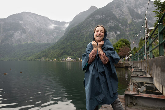 Young woman in a raincoat standing on the shore of a lake in the rain and smiling at the camera, Hallstatt, Austria