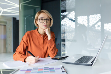 Portrait of young thinking business woman, blonde at workplace inside office concentrating and looking seriously at camera, female boss using laptop behind paper work with documents and reports.