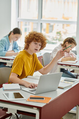 Redhead teen schoolboy holding pencil and using laptop while talking during lesson in classroom
